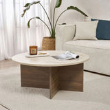 Table basse ronde avec plateau en travertin et base en bois foncé en forme de croix. Banc et plante en arrière-plan.