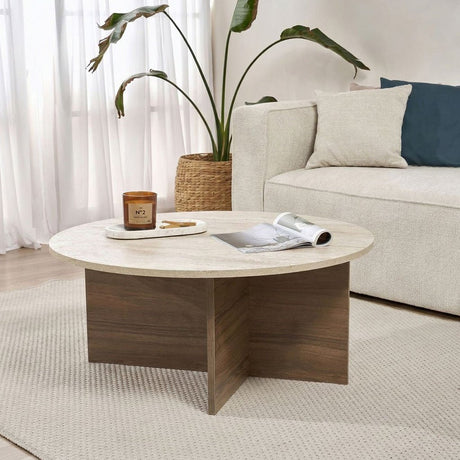 Table basse ronde avec plateau en travertin et base en bois foncé en forme de croix. Banc et plante en arrière-plan.